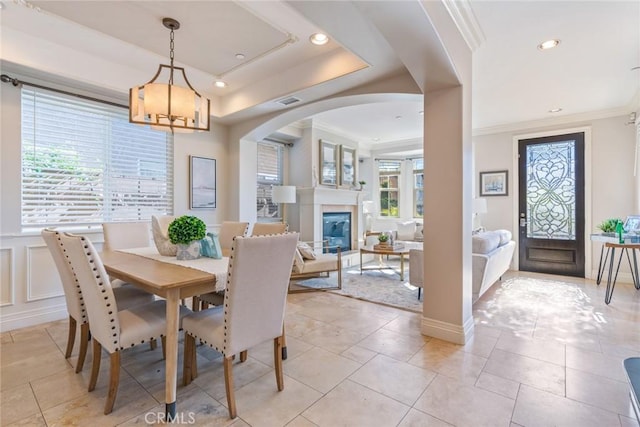 dining area featuring a tray ceiling, ornamental molding, arched walkways, a decorative wall, and a glass covered fireplace