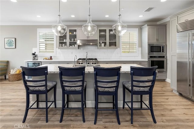 kitchen with visible vents, a center island with sink, under cabinet range hood, built in appliances, and light wood-type flooring