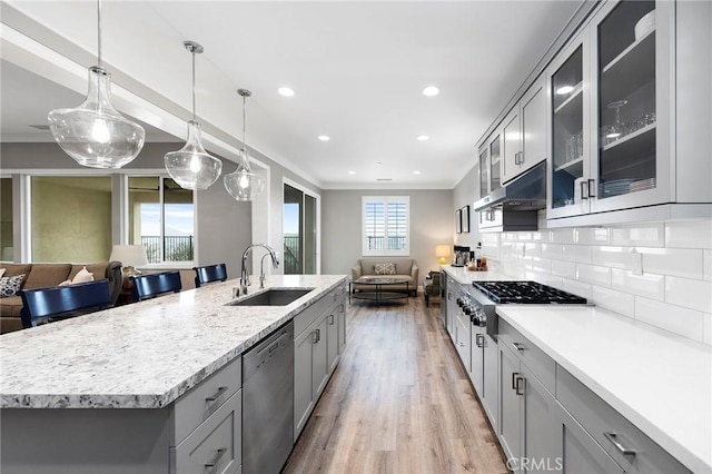 kitchen with a sink, gray cabinetry, under cabinet range hood, appliances with stainless steel finishes, and open floor plan