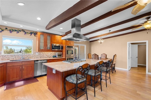 kitchen with ceiling fan, island exhaust hood, brown cabinetry, stainless steel appliances, and a sink