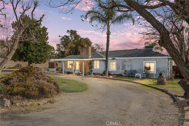 single story home featuring roof with shingles, a chimney, dirt driveway, and stucco siding