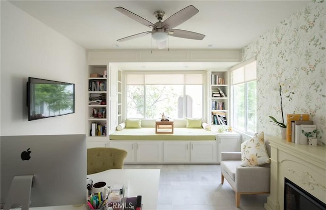 sitting room with built in shelves, a glass covered fireplace, a ceiling fan, and wallpapered walls