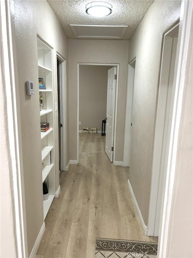 hallway with attic access, built in features, baseboards, a textured ceiling, and light wood-style floors