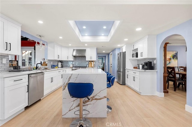 kitchen featuring arched walkways, a raised ceiling, appliances with stainless steel finishes, wall chimney range hood, and light wood-type flooring