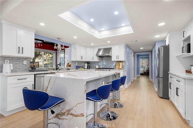 kitchen featuring stainless steel appliances, a breakfast bar, wall chimney range hood, a center island, and a tray ceiling