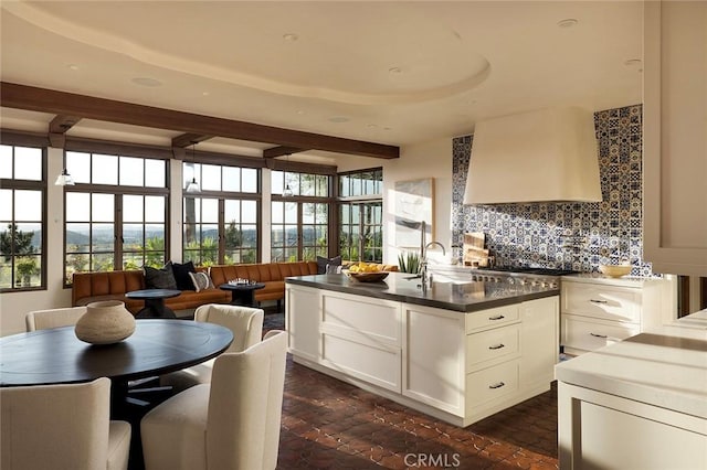 kitchen with ventilation hood, beam ceiling, decorative backsplash, brick floor, and white cabinets