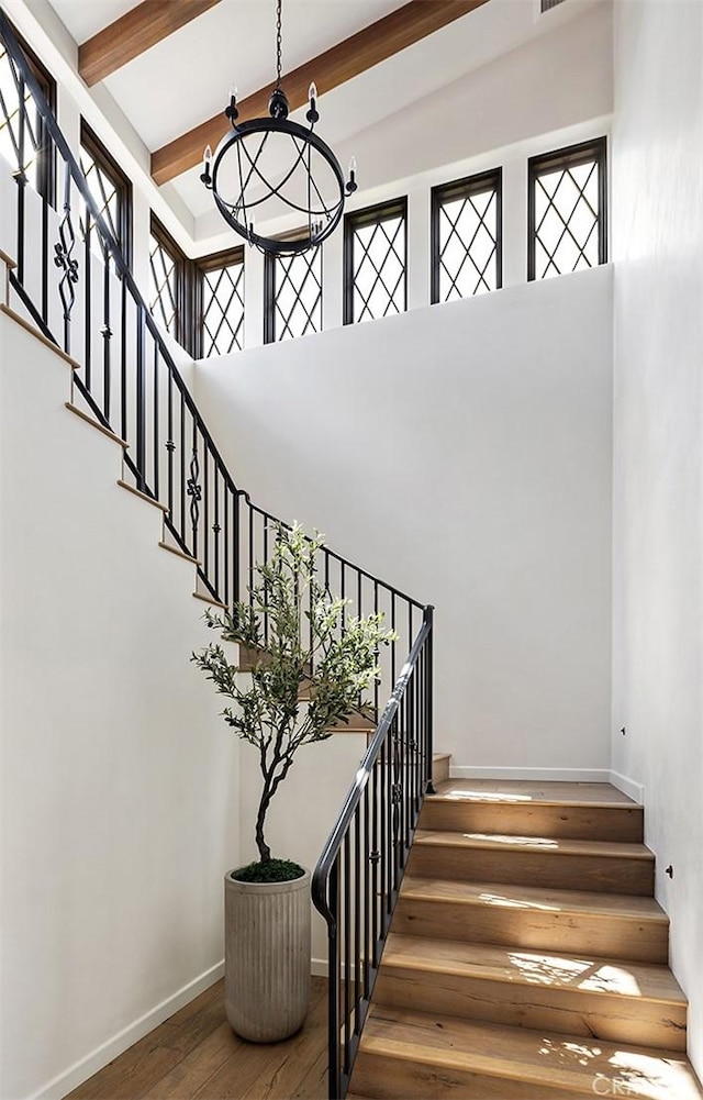 staircase featuring wood finished floors, baseboards, an inviting chandelier, beam ceiling, and a towering ceiling