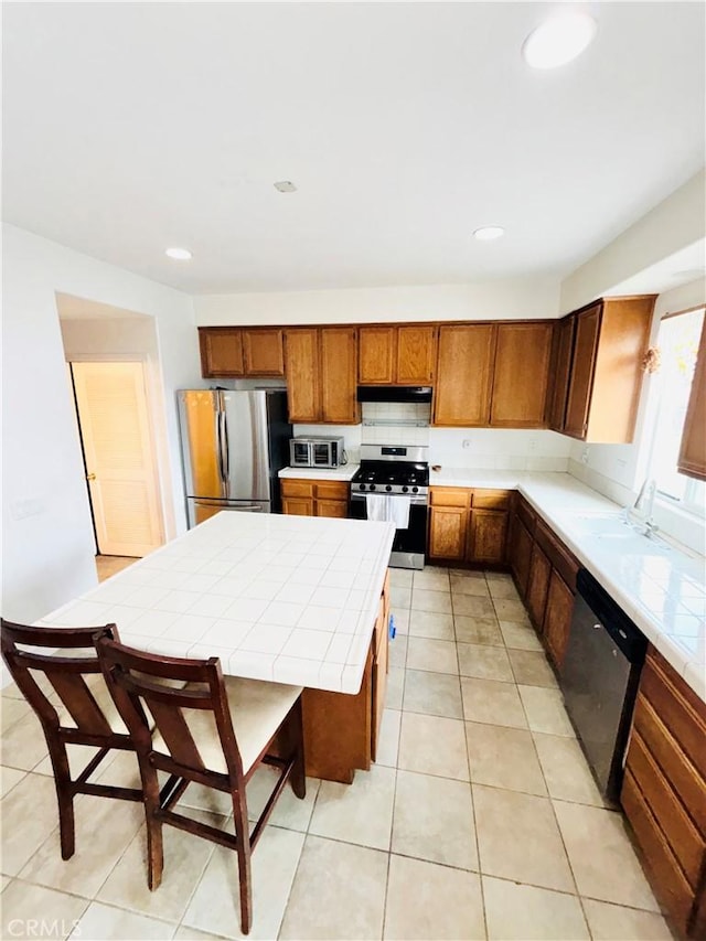 kitchen featuring tile countertops, light tile patterned floors, brown cabinetry, a sink, and stainless steel appliances