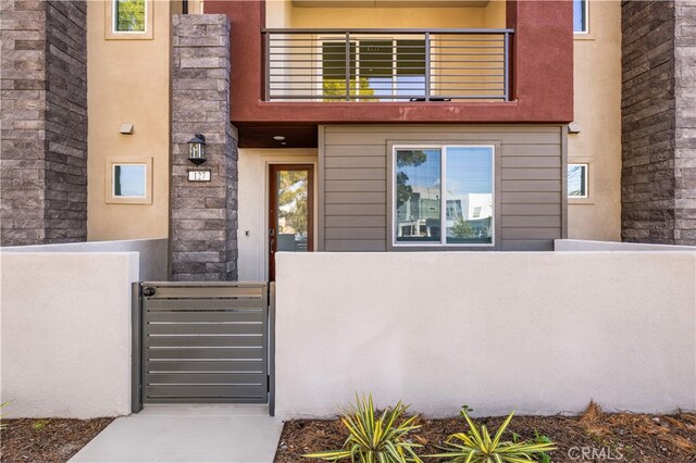 view of exterior entry with fence, stucco siding, stone siding, and a balcony