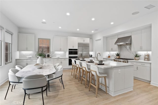 kitchen featuring a center island with sink, double wall oven, a sink, built in refrigerator, and wall chimney range hood