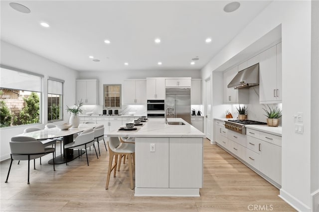 kitchen with wall chimney exhaust hood, a center island with sink, light wood finished floors, and stainless steel appliances