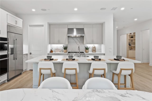 kitchen with backsplash, under cabinet range hood, light wood-type flooring, stainless steel built in fridge, and a kitchen breakfast bar