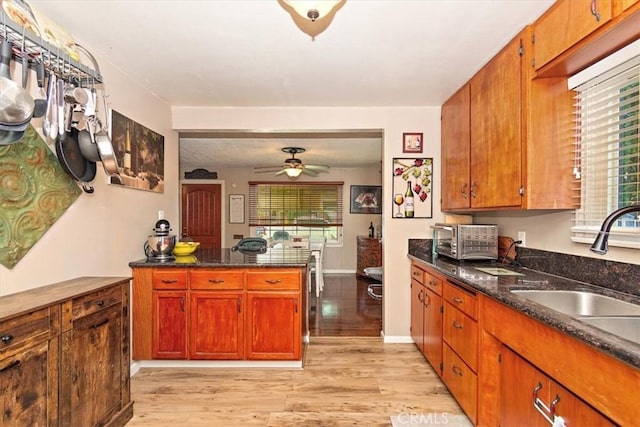 kitchen with brown cabinets, a ceiling fan, dark stone counters, a sink, and light wood-type flooring