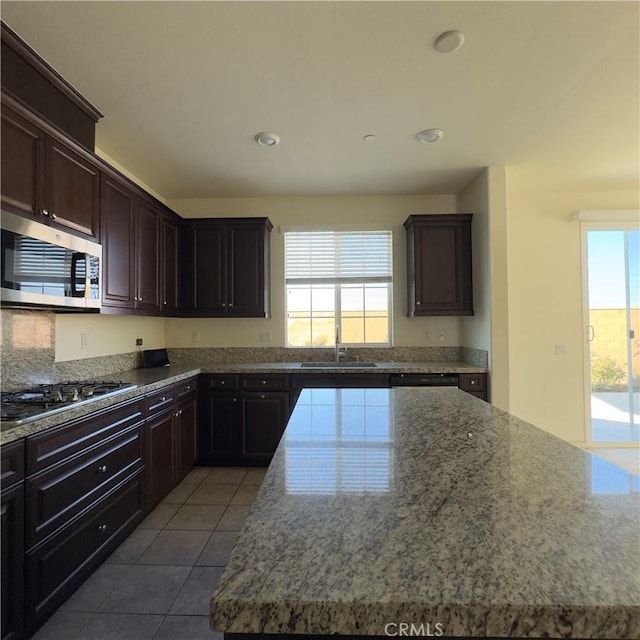 kitchen with appliances with stainless steel finishes, a sink, dark brown cabinetry, light stone countertops, and tile patterned flooring