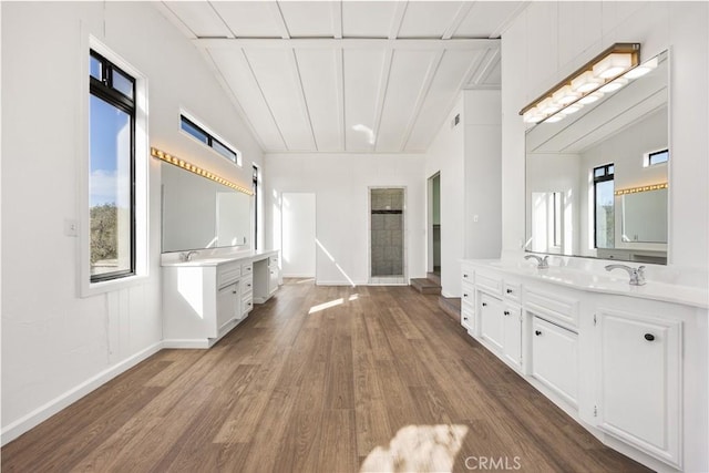 full bath featuring two vanities, wood finished floors, baseboards, vaulted ceiling, and tiled shower