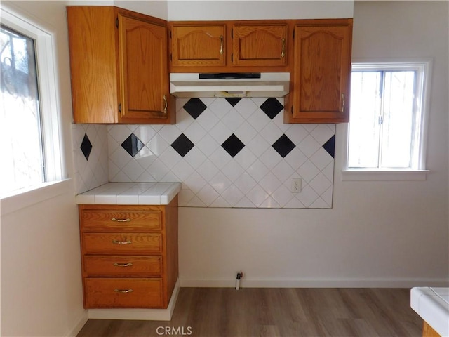 kitchen featuring decorative backsplash, brown cabinetry, light wood-type flooring, under cabinet range hood, and baseboards