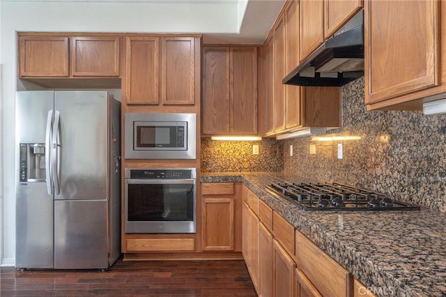 kitchen with dark wood-type flooring, under cabinet range hood, stainless steel appliances, brown cabinetry, and decorative backsplash