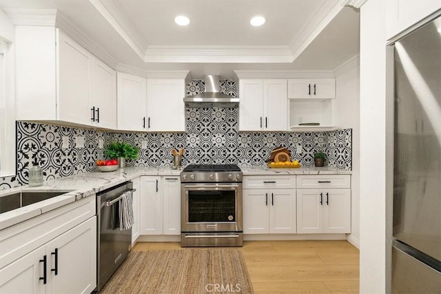 kitchen featuring light wood finished floors, stainless steel appliances, white cabinets, a raised ceiling, and wall chimney exhaust hood