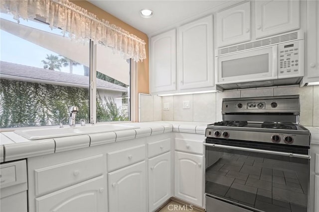 kitchen with white microwave, backsplash, tile counters, stainless steel range with gas stovetop, and white cabinets