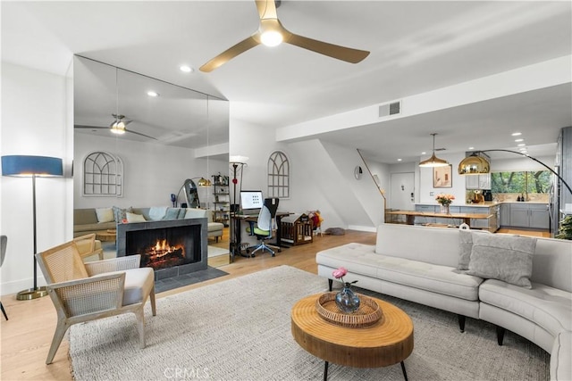 living room featuring light wood-style flooring, a ceiling fan, visible vents, and a warm lit fireplace