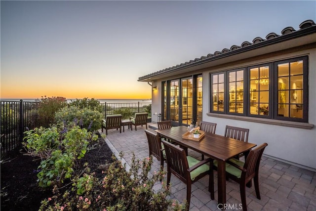 patio terrace at dusk featuring french doors, outdoor dining area, a water view, and fence