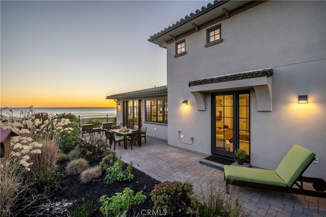 back of house at dusk featuring a water view, fence, stucco siding, outdoor dining area, and a patio area