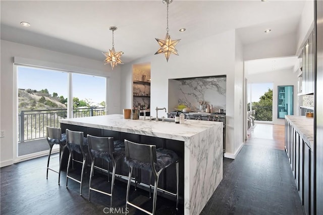 kitchen with light stone counters, dark wood-style floors, baseboards, an island with sink, and decorative light fixtures