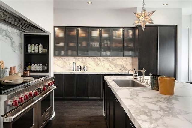 kitchen with light stone counters, a sink, backsplash, range with two ovens, and hanging light fixtures