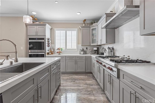 kitchen featuring gray cabinets, a sink, stainless steel appliances, ornamental molding, and wall chimney range hood