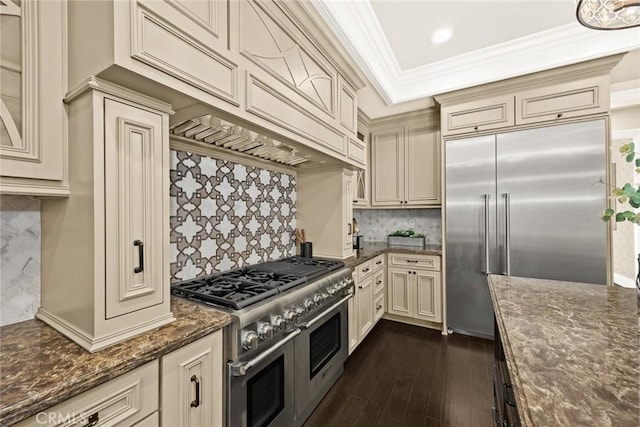 kitchen featuring ornamental molding, premium appliances, dark stone countertops, dark wood-style floors, and cream cabinetry