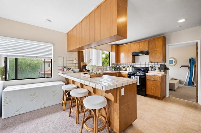 kitchen with tile counters, under cabinet range hood, black gas range, brown cabinets, and a peninsula