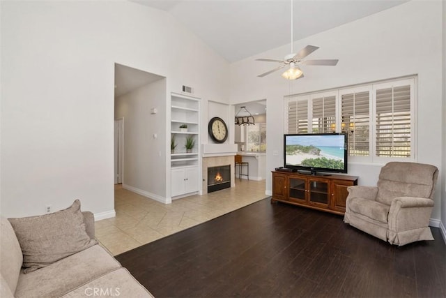 living room with baseboards, ceiling fan, a tiled fireplace, built in features, and wood finished floors