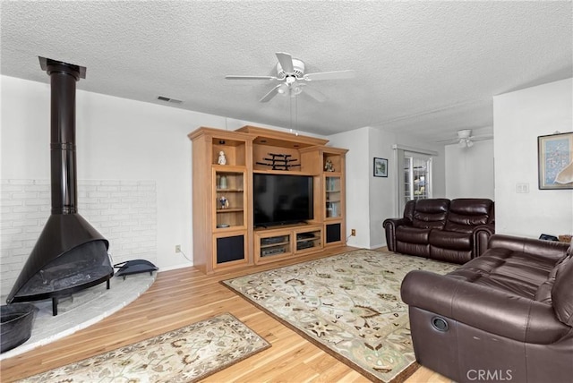 living room featuring visible vents, a ceiling fan, a textured ceiling, wood finished floors, and a wood stove