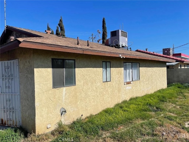 view of side of property featuring central AC unit, stucco siding, and roof with shingles