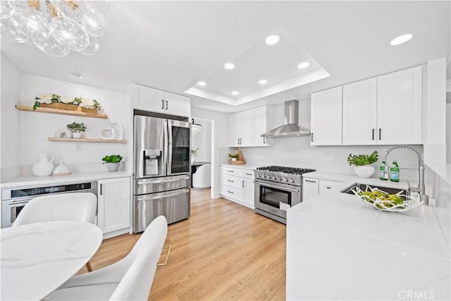 kitchen featuring wall chimney range hood, light wood-style flooring, appliances with stainless steel finishes, a raised ceiling, and a sink