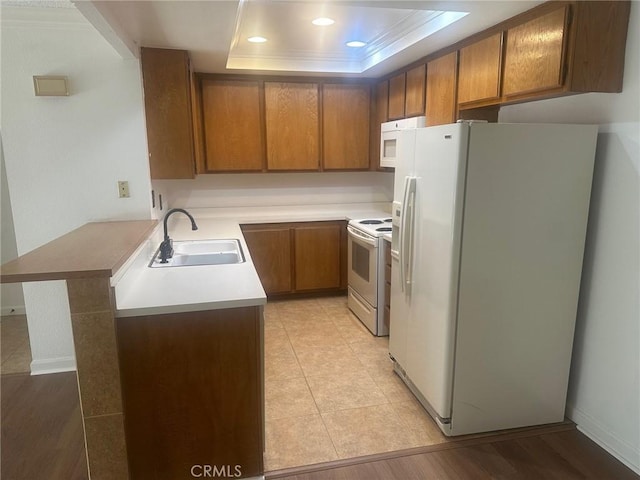 kitchen featuring brown cabinets, a sink, white appliances, a peninsula, and a raised ceiling