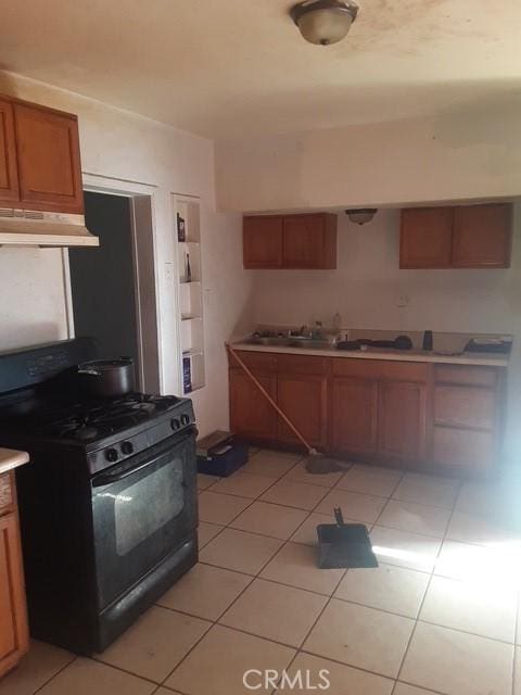 kitchen featuring black gas range, under cabinet range hood, brown cabinetry, light countertops, and light tile patterned floors