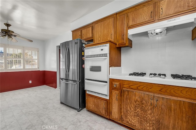 kitchen with a warming drawer, under cabinet range hood, white appliances, brown cabinetry, and light countertops