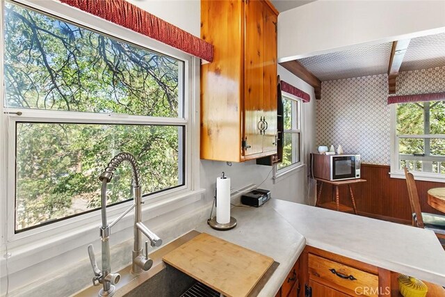 kitchen with a wainscoted wall, wallpapered walls, light countertops, stainless steel microwave, and brown cabinets