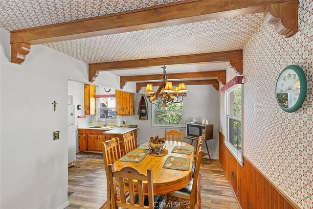 dining area with beamed ceiling, a notable chandelier, light wood-style flooring, and wallpapered walls