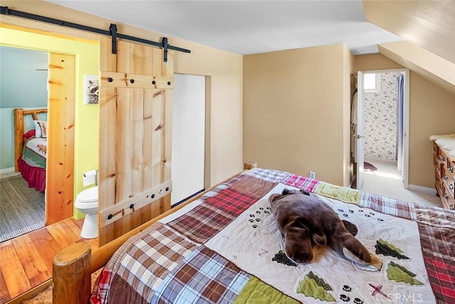 bedroom with a barn door, wood finished floors, ensuite bathroom, and vaulted ceiling