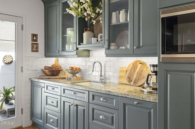 kitchen with glass insert cabinets, light stone countertops, built in microwave, gray cabinets, and a sink