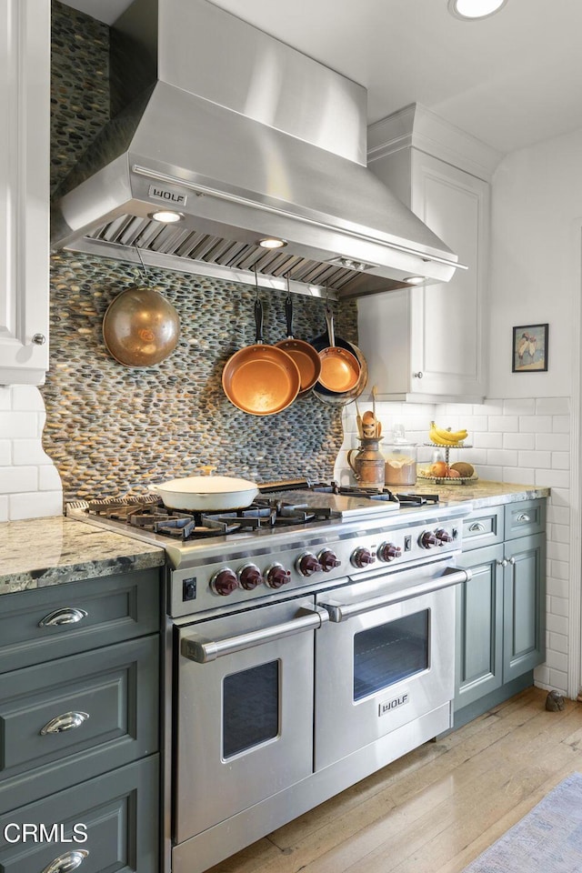 kitchen with light wood finished floors, wall chimney range hood, light stone countertops, range with two ovens, and white cabinets