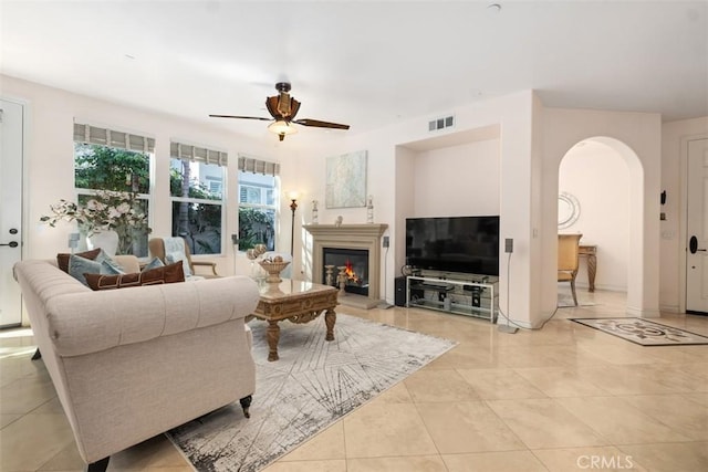 living room featuring visible vents, light tile patterned floors, a warm lit fireplace, arched walkways, and a ceiling fan