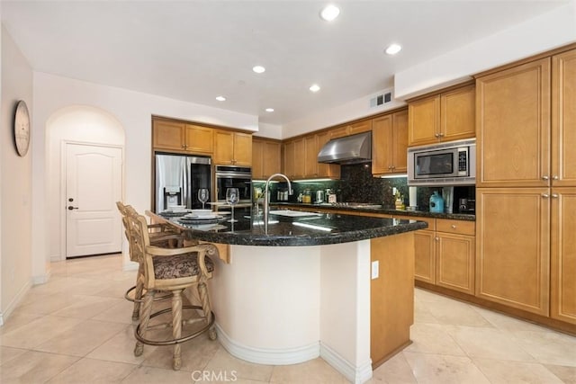 kitchen with a sink, wall chimney range hood, brown cabinets, appliances with stainless steel finishes, and a kitchen island with sink