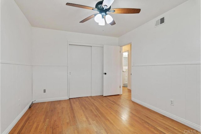 unfurnished bedroom featuring visible vents, a wainscoted wall, light wood-type flooring, a closet, and a ceiling fan