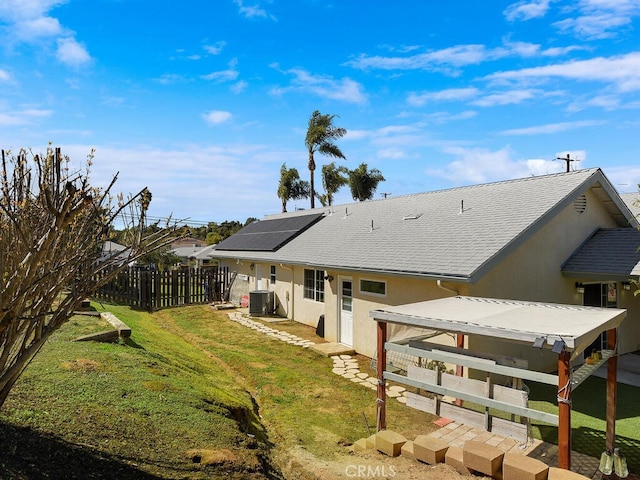 back of property with stucco siding, fence, central AC unit, a yard, and roof mounted solar panels