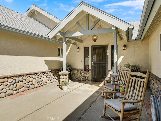 doorway to property featuring a shingled roof, stucco siding, covered porch, and stone siding