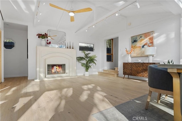 living room with track lighting, a ceiling fan, a glass covered fireplace, wood finished floors, and vaulted ceiling with beams