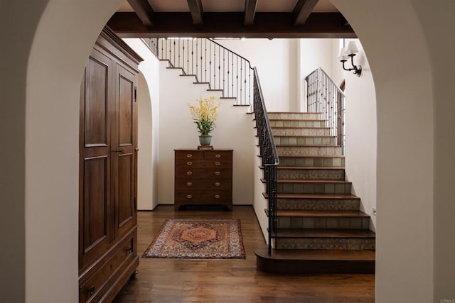 staircase with arched walkways, beam ceiling, and wood finished floors